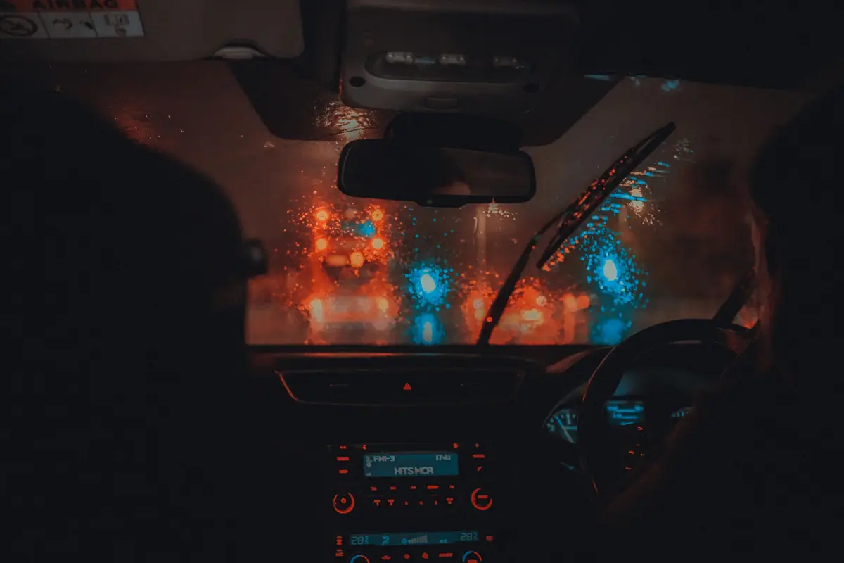 Rückspiegel hinter regennasser Windschutzscheibe, dahinter rote und blaue Lichter der Straße